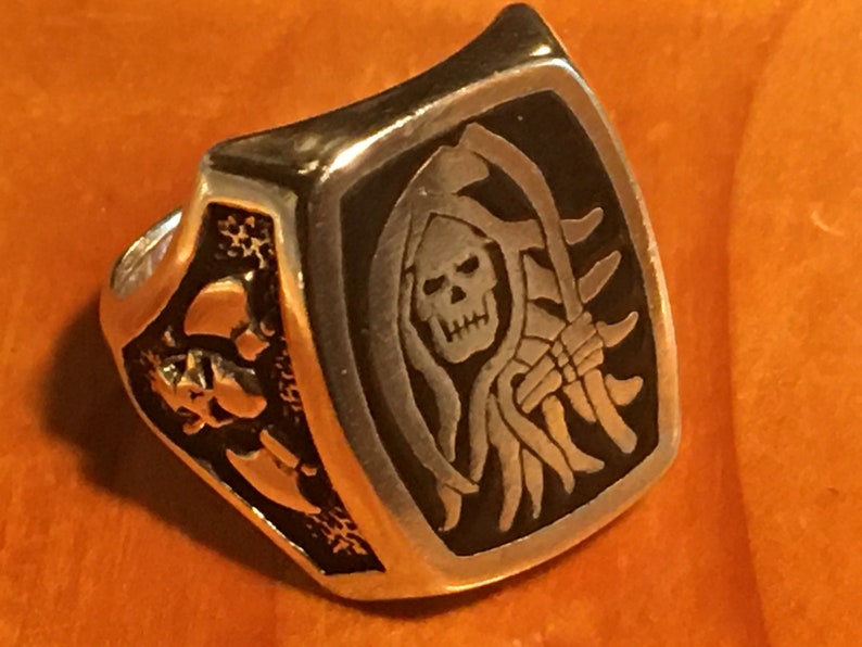 May include: A silver-colored ring with a rectangular face featuring a Grim Reaper design. The ring's face has a black background with a detailed engraving of the Grim Reaper holding a scythe. The sides of the ring feature skull designs.
