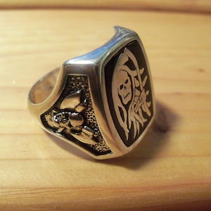 May include: Silver-toned ring with a rectangular black face featuring a white Grim Reaper design. The ring has a detailed floral pattern on the side and is set against a wooden surface. A bold, statement piece.