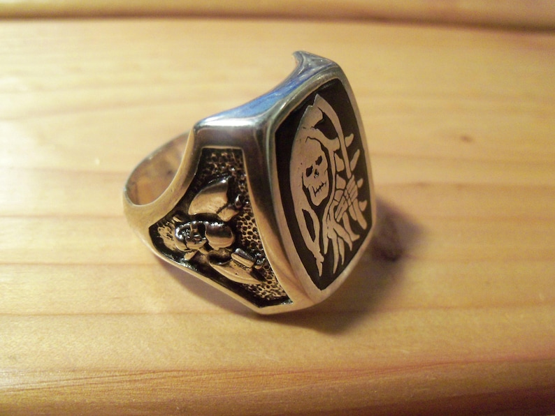 May include: Silver-toned ring with a rectangular black face featuring a white Grim Reaper design. The ring has a detailed floral pattern on the side and is set against a wooden surface. A bold, statement piece.