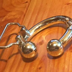 May include: A silver belt buckle with a unique design featuring a curved, rounded shape with two ball-shaped ends. The buckle has a simple, rectangular loop for attaching the belt.