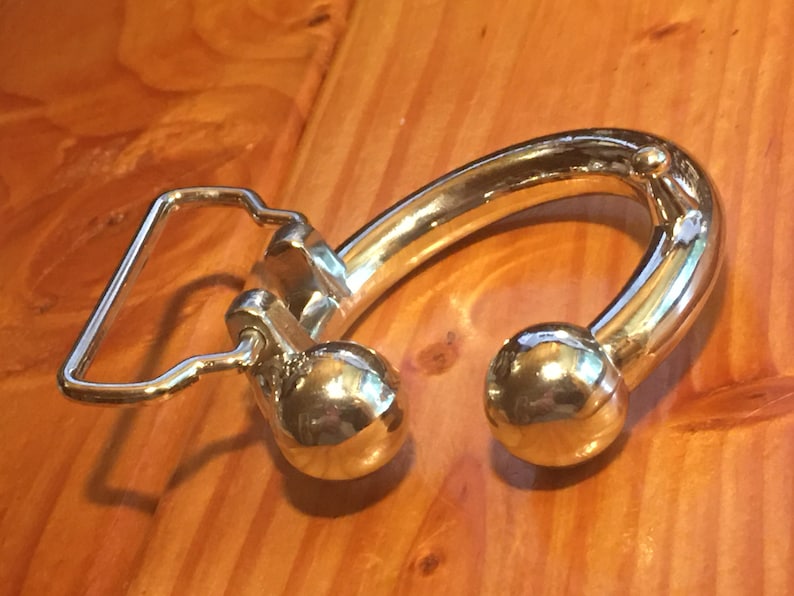 May include: A silver belt buckle with a unique design featuring a curved, rounded shape with two ball-shaped ends. The buckle has a simple, rectangular loop for attaching the belt.