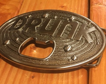 Może przedstawiać: Srebrna metalowa klamra do paska z otwieraczem do butelek. Klamra ma owalny kształt i ma wytłoczone słowo "DRUNK".