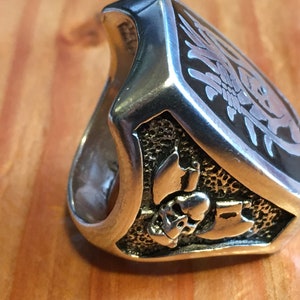 May include: A silver-toned ring with a detailed design. The ring features a shield-shaped face with an etched floral pattern and a skull with crossed axes. The ring is set against a wooden surface.