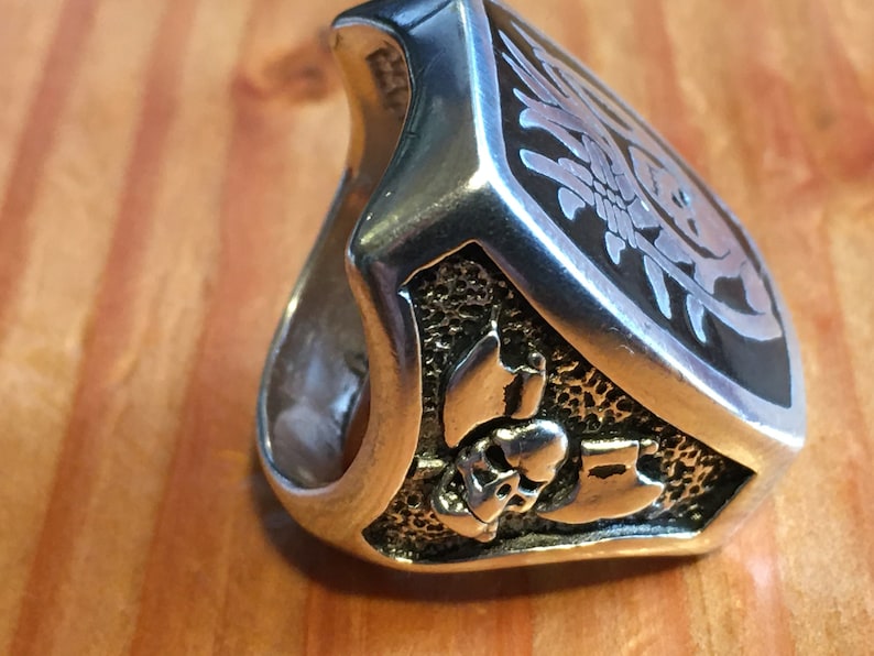 May include: A silver-toned ring with a detailed design. The ring features a shield-shaped face with an etched floral pattern and a skull with crossed axes. The ring is set against a wooden surface.