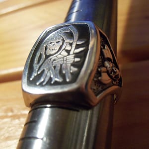 May include: A silver-colored ring featuring a detailed grim reaper design with a scythe. The ring has a rectangular face with a black background, and is displayed on a metal ring sizer. The ring's design is intricate and detailed.