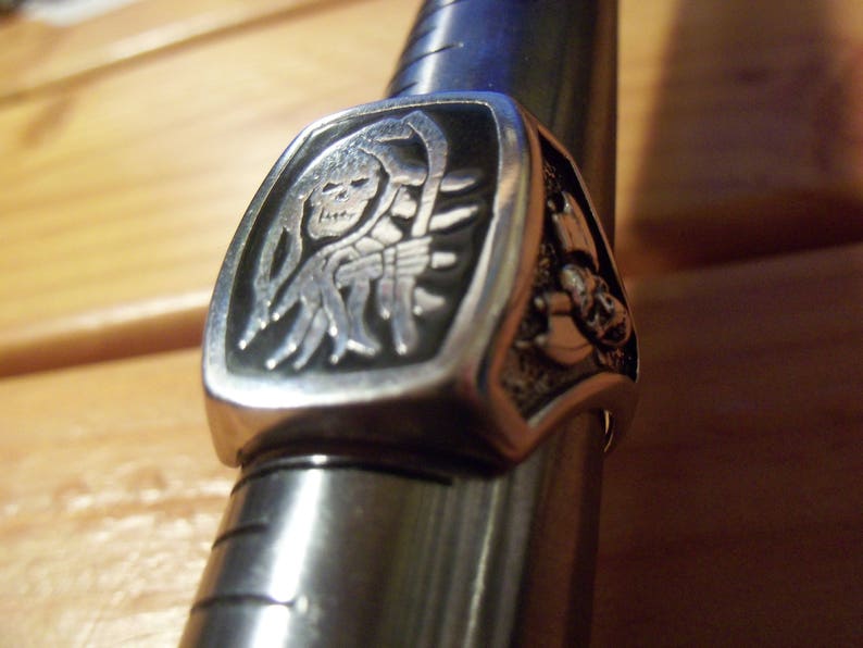 May include: A silver-colored ring featuring a detailed grim reaper design with a scythe. The ring has a rectangular face with a black background, and is displayed on a metal ring sizer. The ring's design is intricate and detailed.