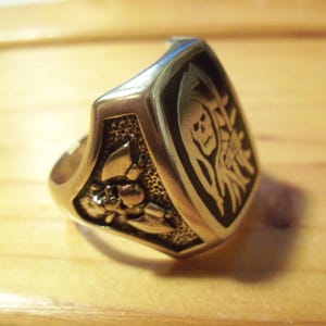May include: Silver-toned ring with a shield-shaped face. The face features a black background with a Grim Reaper design and a floral motif on the side. The ring is likely made of metal.
