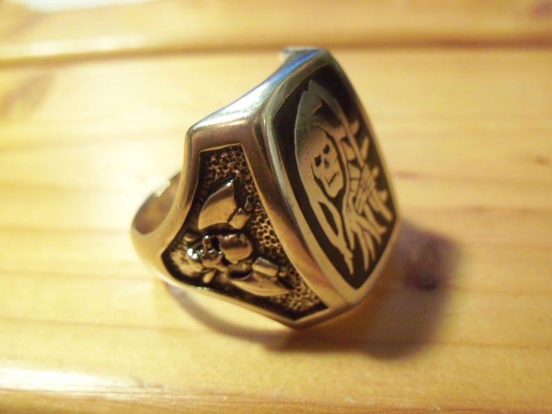 May include: Silver-toned ring with a shield-shaped face. The face features a black background with a Grim Reaper design and a floral motif on the side. The ring is likely made of metal.