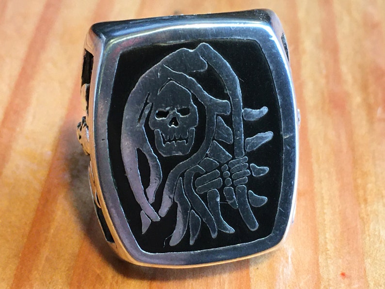 May include: Silver-toned ring featuring a black rectangular face with a detailed Grim Reaper design. The reaper is depicted with a skull and holding a scythe. The ring has a square shape and a detailed band.