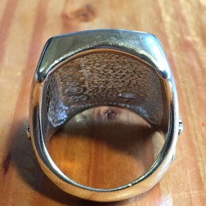May include: A gold-colored ring with a rectangular top and rounded edges. The inside of the ring has a textured pattern and the numbers "2008" and "89" are visible. The ring is set against a wooden surface.