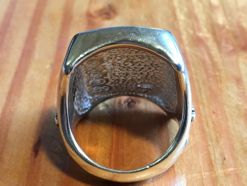 May include: A gold-colored ring with a rectangular top and rounded edges. The inside of the ring has a textured pattern and the numbers "2008" and "89" are visible. The ring is set against a wooden surface.