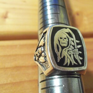 May include: Silver-toned ring featuring a rectangular face with a black background and a silver Grim Reaper design. The ring is on a metal ring sizer. The side of the ring has a decorative skull design.