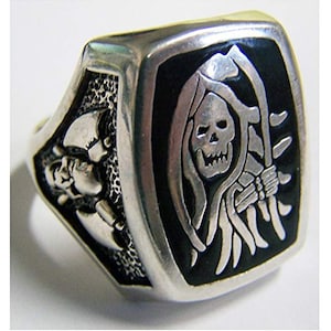 May include: A silver-toned ring with a rectangular face featuring a grim reaper design. The reaper is depicted with a skull face, holding a scythe against a black background. The ring's sides have skull and bone details.