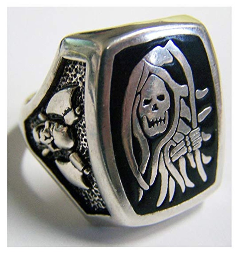 May include: A silver-toned ring with a rectangular face featuring a grim reaper design. The reaper is depicted with a skull face, holding a scythe against a black background. The ring's sides have skull and bone details.