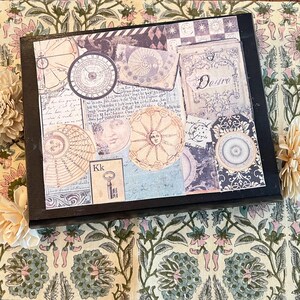 Puede incluir: Una caja decorativa con marco negro con un collage de imágenes de estilo vintage, que incluyen brújulas, cuerpos celestes y texto manuscrito. La palabra "Desire" es visible. La caja está sobre una superficie con estampado floral.