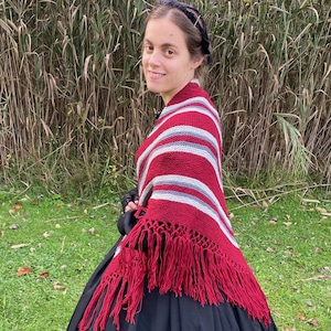 Puede incluir: Una persona lleva un vestido negro y un chal a rayas rojas, blancas y grises con flecos rojos. El chal está drapeado sobre los hombros. El fondo presenta hierba alta y follaje verde.