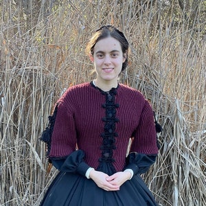 Peut inclure: Une femme portant un pull en tricot bordeaux avec des bordures noires et une jupe noire. Elle se tient dans un champ d'herbe haute.