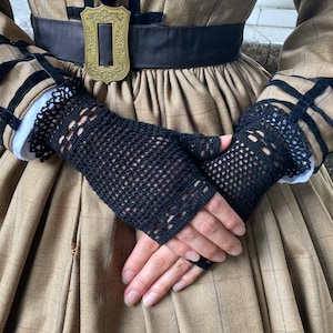 Puede incluir: Guantes sin dedos de crochet negro con un delicado dise&ntilde;o calado. Los guantes se usan sobre un pu&ntilde;o blanco y se combinan con un vestido a cuadros marr&oacute;n. Los guantes son un accesorio elegante.