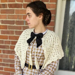 Puede incluir: Una mujer con un chal de ganchillo color crema sobre un vestido a cuadros marrón y blanco. Lleva un lazo negro, guantes negros y un cinturón negro. El conjunto tiene una estética vintage.