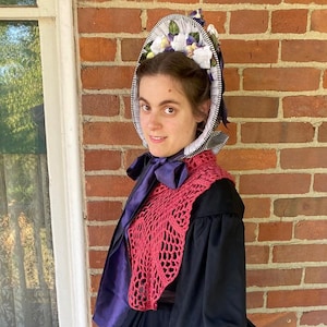 May include: A woman wearing a black dress with a large skirt, a red crocheted shawl, and a white bonnet with purple flowers.