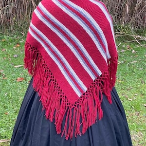 Puede incluir: Una persona con un chal a rayas rojas y blancas con flecos rojos, una gorra azul oscuro y una falda larga negra. El chal tiene un patr&oacute;n de rayas diagonales. La persona est&aacute; de pie en una zona cubierta de hierba con hierba alta al fondo.
