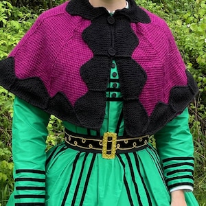 Peut inclure: Une capelette tricotée magenta et noire avec un col noir et une fermeture boutonnée. La capelette est portée sur une robe verte avec des rayures verticales noires et une ceinture à boucle dorée. Les manches ont des rayures noires.