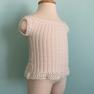 May include: A white crocheted top with a delicate lace trim. The top has a scoop neck and short sleeves. It is displayed on a white mannequin.