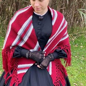 Puede incluir: Un chal a rayas rojas, blancas y grises con flecos. El chal se lleva sobre un vestido negro. La imagen tambi&eacute;n muestra guantes de encaje negros y un gorro negro. El chal es un accesorio acogedor.