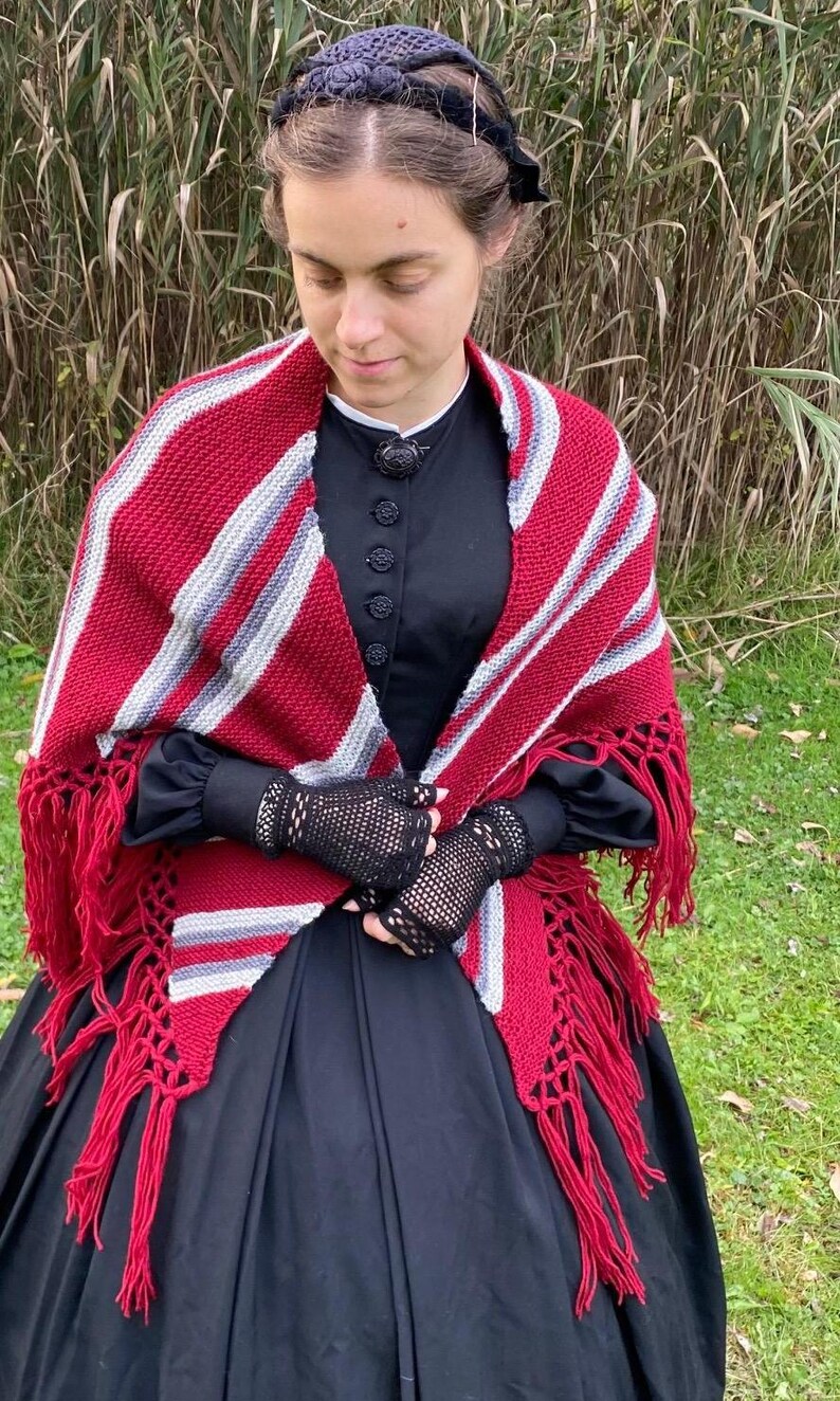 Puede incluir: Un chal a rayas rojas, blancas y grises con flecos. El chal se lleva sobre un vestido negro. La imagen tambi&eacute;n muestra guantes de encaje negros y un gorro negro. El chal es un accesorio acogedor.