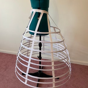 May include: A white, circular, multi-tiered cage crinoline with a white ribbon waistband. The crinoline is on a dress form.