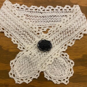 May include: A white, crocheted collar with a decorative black button. The collar is made of delicate, open-weave crochet and features a scalloped edge. The black button is round with an ornate design.