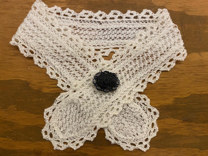May include: A white, crocheted collar with a decorative black button. The collar is made of delicate, open-weave crochet and features a scalloped edge. The black button is round with an ornate design.