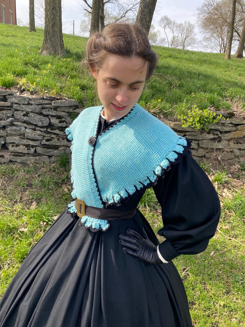 May include: A vintage outfit comprising a light blue crocheted shawl with black edging and a black dress. The shawl has a scalloped edge and a decorative button. The dress features long sleeves, a wide skirt, a black belt, and a gold buckle.