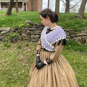 May include: A woman in a tan and brown plaid dress with a full skirt, black gloves, and a lavender shawl with white edging. The dress features long sleeves with black and white detailing. Her hair is styled in a braided updo.