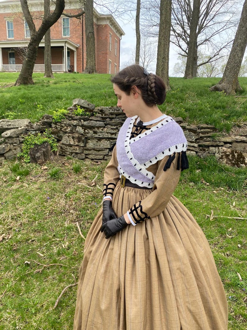 May include: A woman in a tan and brown plaid dress with a full skirt, black gloves, and a lavender shawl with white edging. The dress features long sleeves with black and white detailing. Her hair is styled in a braided updo.
