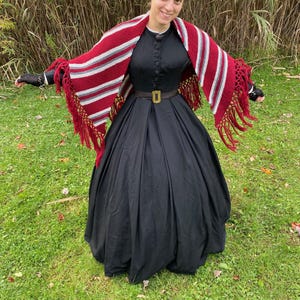 Puede incluir: Una mujer con un vestido negro con cuello alto y detalles de botones. Un chal a rayas rojas, blancas y grises con flecos est&aacute; drapeado sobre los hombros. Un cintur&oacute;n marr&oacute;n con hebilla dorada ci&ntilde;e la cintura. El fondo es hierba verde y hierba alta.