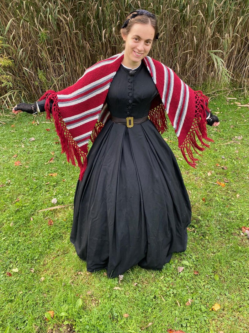 Puede incluir: Una mujer con un vestido negro con cuello alto y detalles de botones. Un chal a rayas rojas, blancas y grises con flecos est&aacute; drapeado sobre los hombros. Un cintur&oacute;n marr&oacute;n con hebilla dorada ci&ntilde;e la cintura. El fondo es hierba verde y hierba alta.