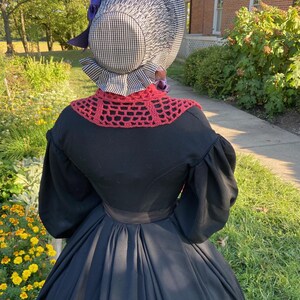 K&ouml;nnte beinhalten: Ein schwarzes Kleid mit weitem Rock und langen &Auml;rmeln, kombiniert mit einer karierten Haube und einem roten geh&auml;kelten Kragen. Die Haube ist mit lila B&auml;ndern und Blumen verziert. Das Outfit erinnert an historische Kleidung.