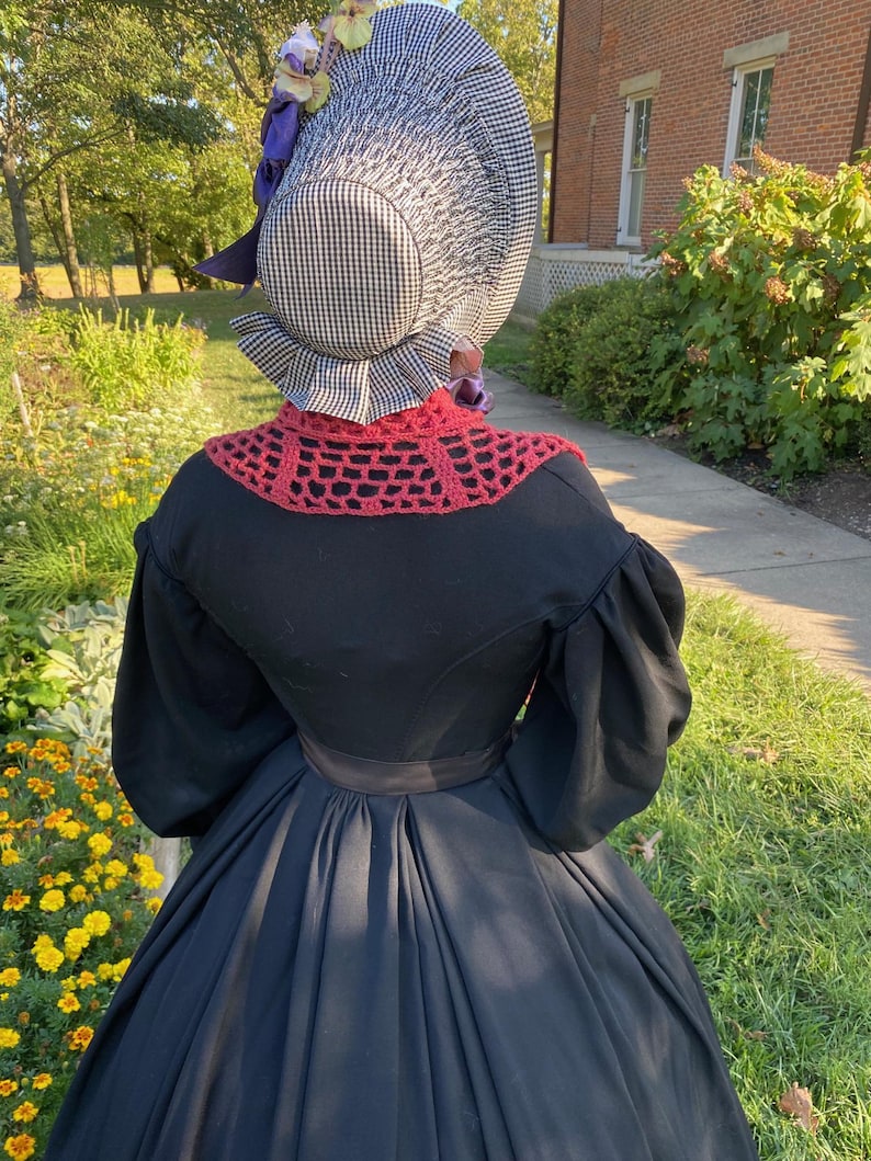 K&ouml;nnte beinhalten: Ein schwarzes Kleid mit weitem Rock und langen &Auml;rmeln, kombiniert mit einer karierten Haube und einem roten geh&auml;kelten Kragen. Die Haube ist mit lila B&auml;ndern und Blumen verziert. Das Outfit erinnert an historische Kleidung.