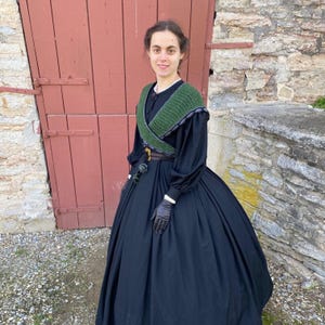 May include: A black Victorian dress with a full skirt and long sleeves. The dress has a green crocheted shawl and black gloves. The person is standing in front of a red door and a stone wall.