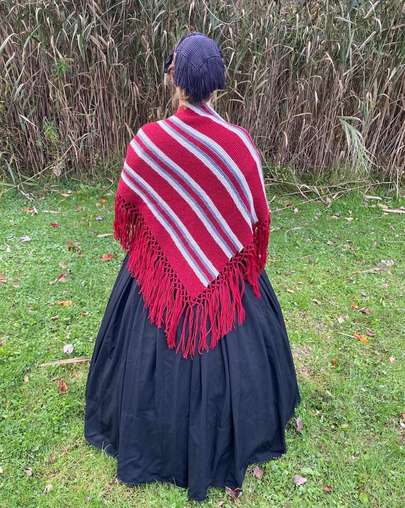 Puede incluir: Una persona con un chal a rayas rojas y blancas con flecos rojos, una gorra azul oscuro y una falda larga negra. El chal tiene un patr&oacute;n de rayas diagonales. La persona est&aacute; de pie en una zona cubierta de hierba con hierba alta al fondo.