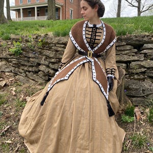 May include: A tan-coloured historical dress with a brown and white crocheted shawl and trim. The dress features a full skirt, a fitted bodice, and a black belt with a gold buckle. Black tassels hang from the shawl.