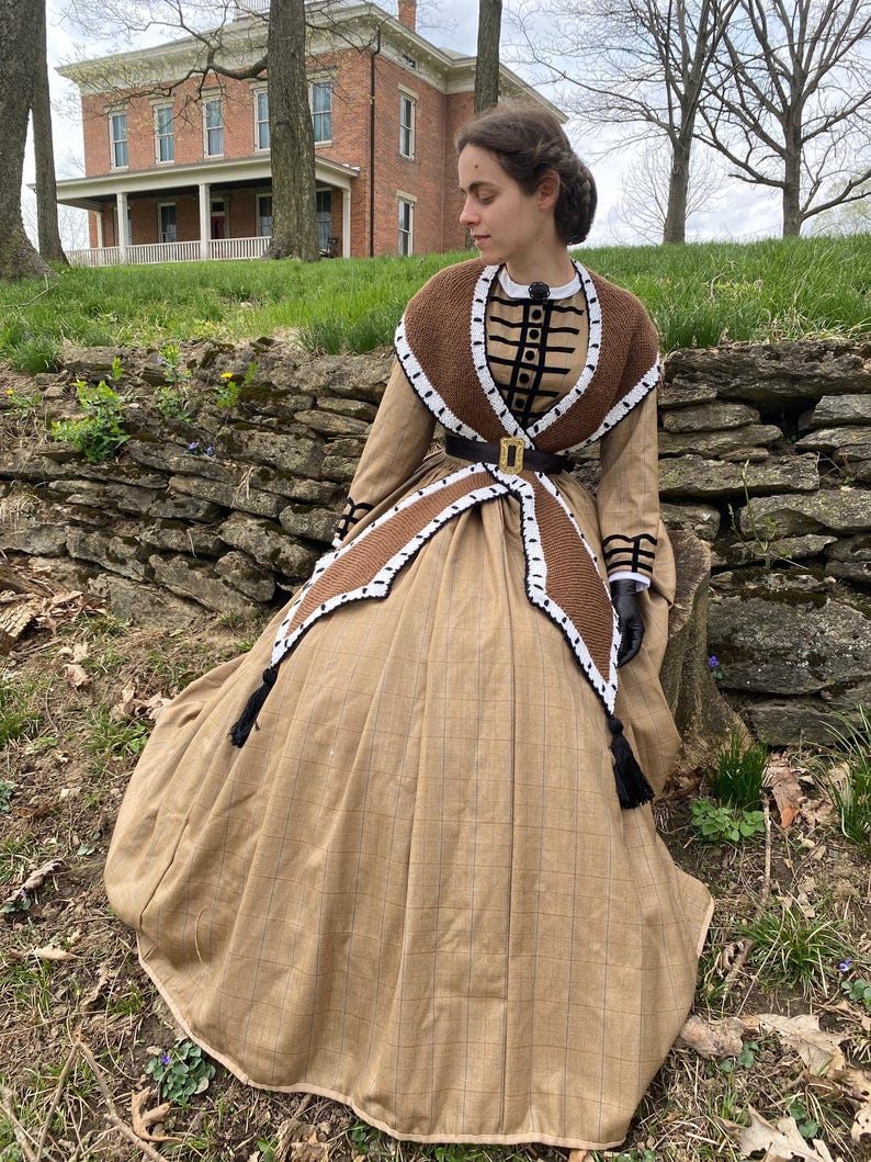 May include: A tan-coloured historical dress with a brown and white crocheted shawl and trim. The dress features a full skirt, a fitted bodice, and a black belt with a gold buckle. Black tassels hang from the shawl.