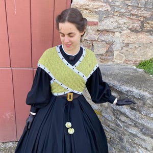 May include: A person wearing a black dress with a long skirt and a green and white knitted top. The top has a geometric design. The outfit is accessorized with a brown belt with a gold buckle and black gloves.