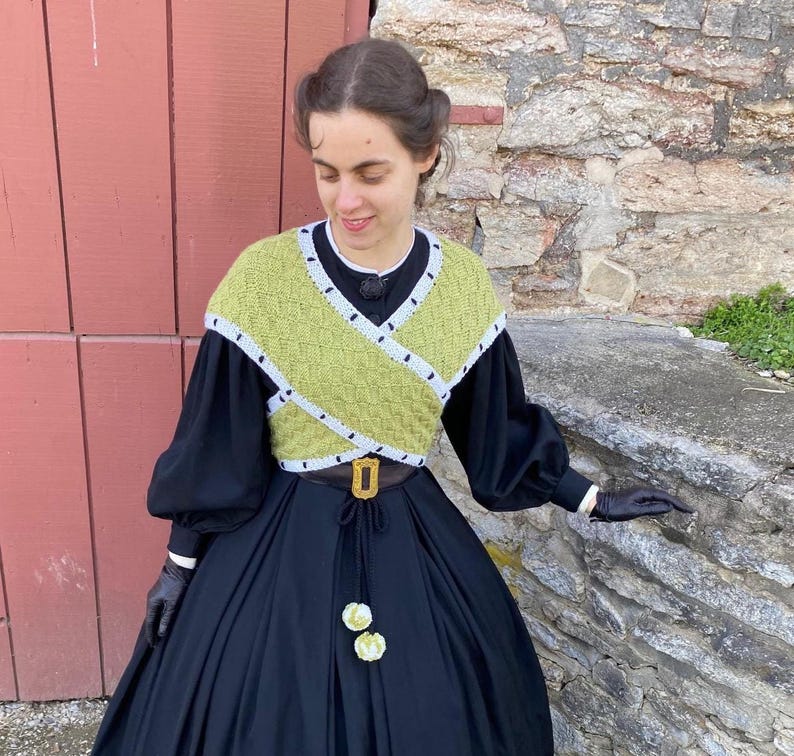 May include: A person wearing a black dress with a long skirt and a green and white knitted top. The top has a geometric design. The outfit is accessorized with a brown belt with a gold buckle and black gloves.