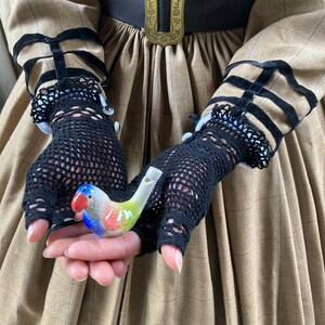 Puede incluir: Un silbato de p&aacute;jaro de cer&aacute;mica de colores, sostenido en guantes de malla negros. El p&aacute;jaro tiene detalles en rojo, azul, verde y amarillo. Los guantes forman parte de un vestido marr&oacute;n con ribetes negros y una hebilla decorativa.