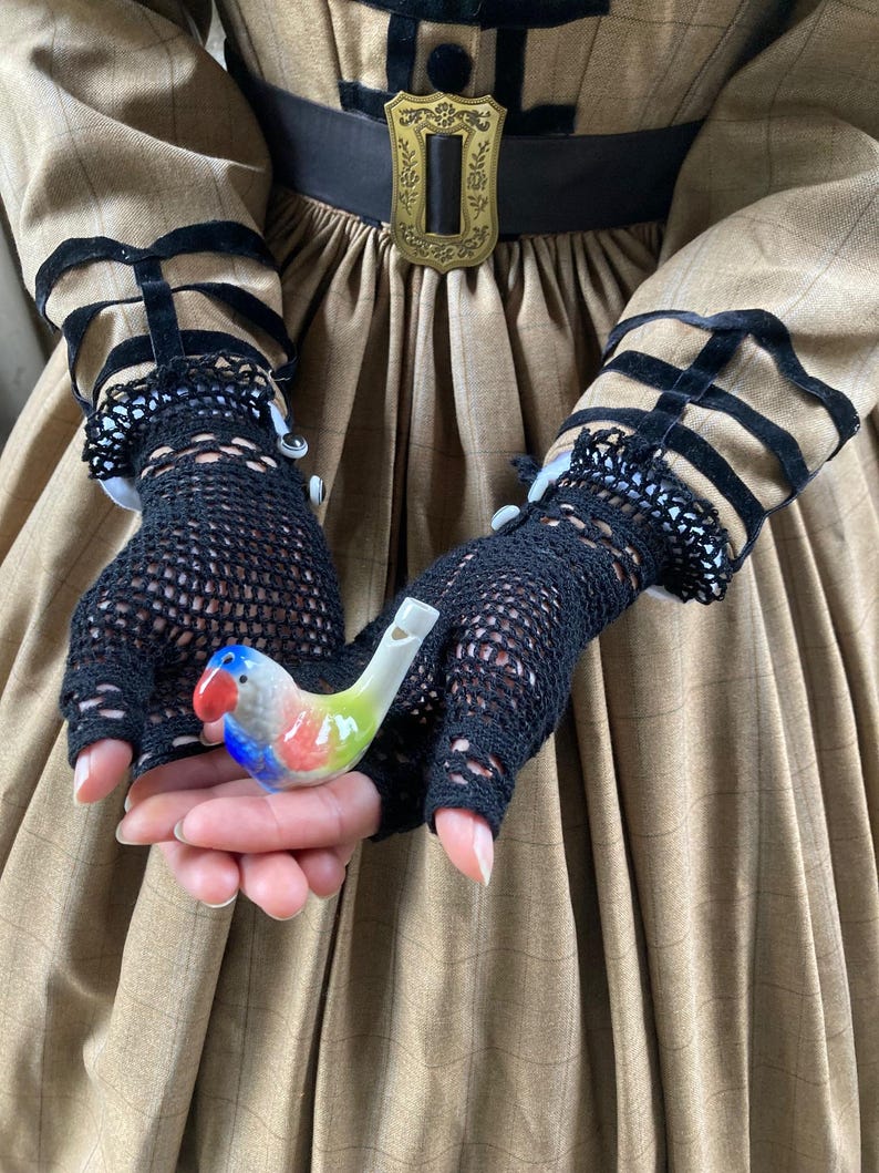 Puede incluir: Un silbato de p&aacute;jaro de cer&aacute;mica de colores, sostenido en guantes de malla negros. El p&aacute;jaro tiene detalles en rojo, azul, verde y amarillo. Los guantes forman parte de un vestido marr&oacute;n con ribetes negros y una hebilla decorativa.