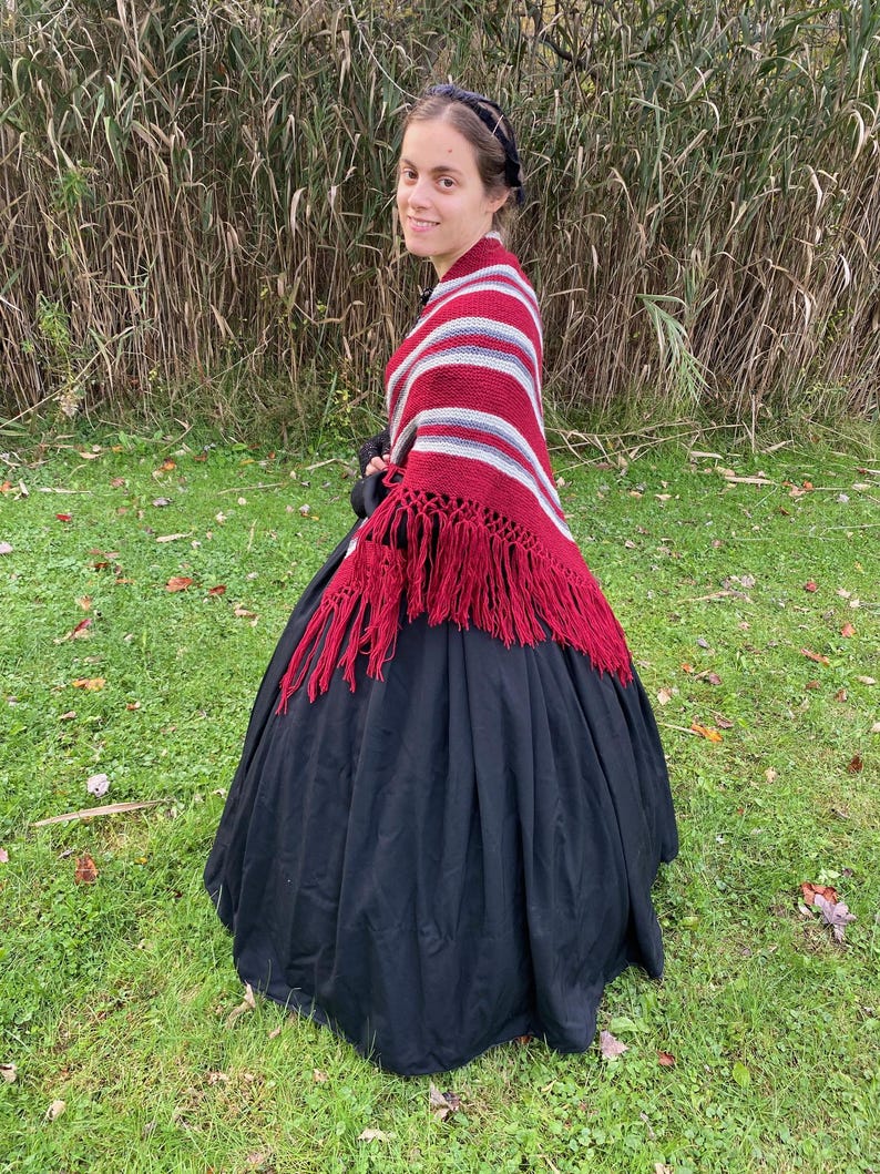 Puede incluir: Una persona lleva un vestido negro y un chal a rayas rojas, blancas y grises con flecos rojos. El chal est&aacute; drapeado sobre los hombros. El fondo presenta hierba alta y follaje verde.