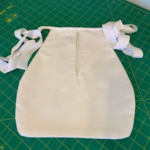 Op de afbeelding: Witte stoffen schort met witte elastische banden. De schort heeft een naad in het midden en een kleine opening aan de bovenkant.