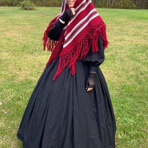 Puede incluir: Una persona vestida con un vestido negro y un chal rojo, blanco y gris con flecos. El chal est&aacute; drapeado sobre la cabeza y los hombros. El vestido tiene mangas largas y una falda amplia. Se usan guantes de encaje negros.